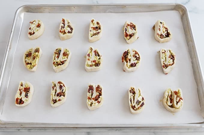 Sur une plaque de cuisson recouverte de papier sulfurisé, des tranches de palmiers feuilletes crus garnies de pesto, tomates séchées, fromage feta et olives