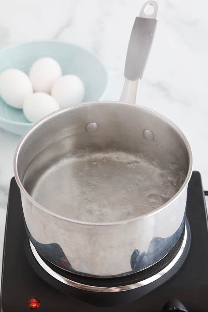 Casserole d'eau en train de bouillir sur une plaque chauffante. 4 gros oeufs dans un bol bleu.